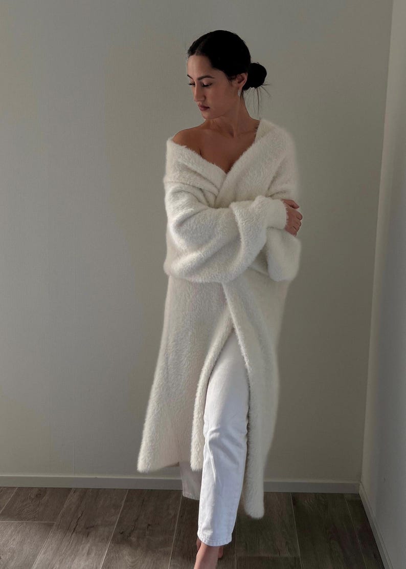 Op de afbeelding: Een lange, witte, pluche cardigan met een diepe V-hals en lange mouwen. De cardigan is open aan de voorkant en het model draagt ​​hem over een witte broek.