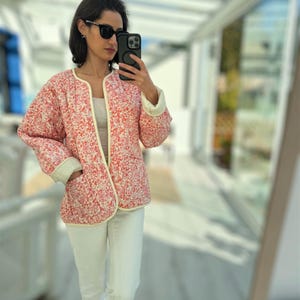 以下が含まれることがあります： 赤と白の花柄のキルティングジャケット。クリーム色のトリムと袖口が付いています。白いジーンズとサングラスを着用しています。ジャケットは開いており、白いトップスが見えます。花柄のデザインが特徴的です。