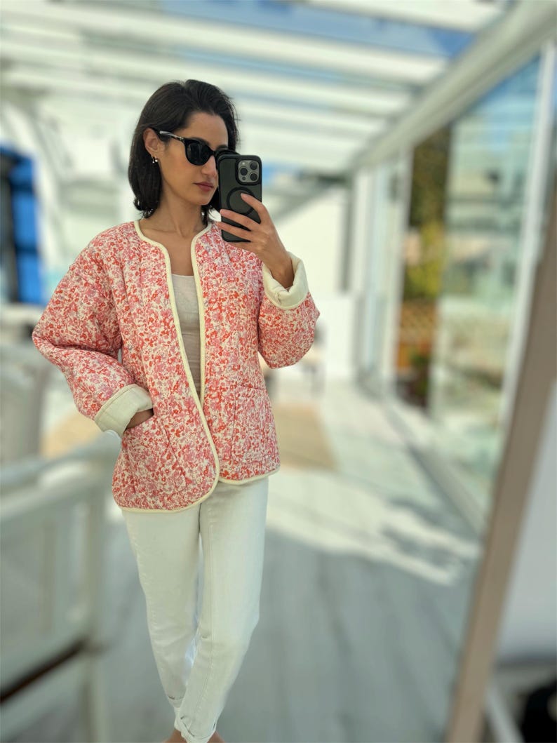 以下が含まれることがあります： 赤と白の花柄のキルティングジャケット。クリーム色のトリムと袖口が付いています。白いジーンズとサングラスを着用しています。ジャケットは開いており、白いトップスが見えます。花柄のデザインが特徴的です。