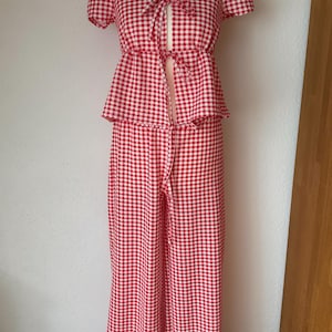 May include: A two-piece outfit featuring a red and white gingham pattern. The top has short puff sleeves, a scoop neckline, and a front tie closure. The matching wide-leg trousers have a high waist and a long, flowing silhouette that reaches the floor.