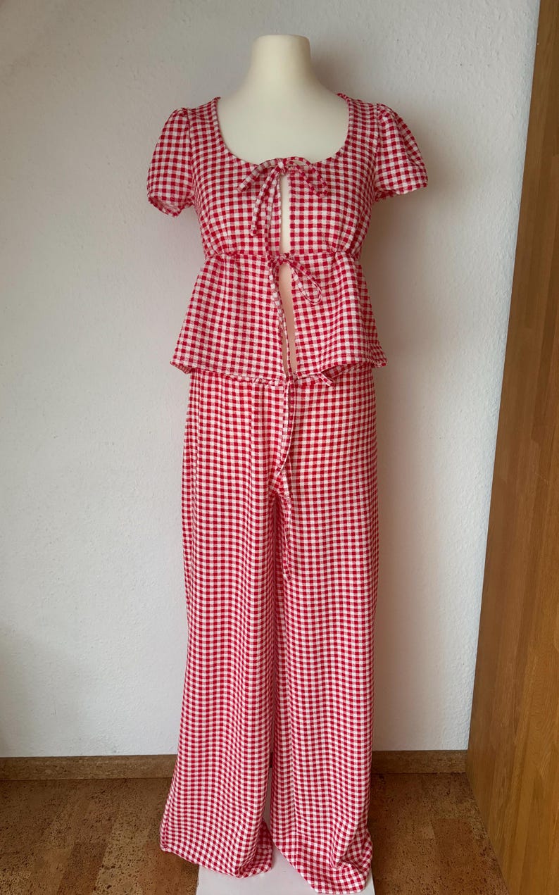 May include: A two-piece outfit featuring a red and white gingham pattern. The top has short puff sleeves, a scoop neckline, and a front tie closure. The matching wide-leg trousers have a high waist and a long, flowing silhouette that reaches the floor.
