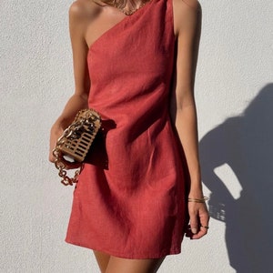 May include: A one-shoulder, short, red linen dress with a fitted silhouette. The dress is sleeveless and has a draped neckline.