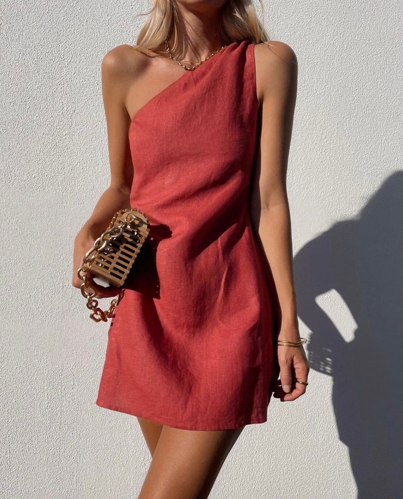 May include: A one-shoulder, short, red linen dress with a fitted silhouette. The dress is sleeveless and has a draped neckline.