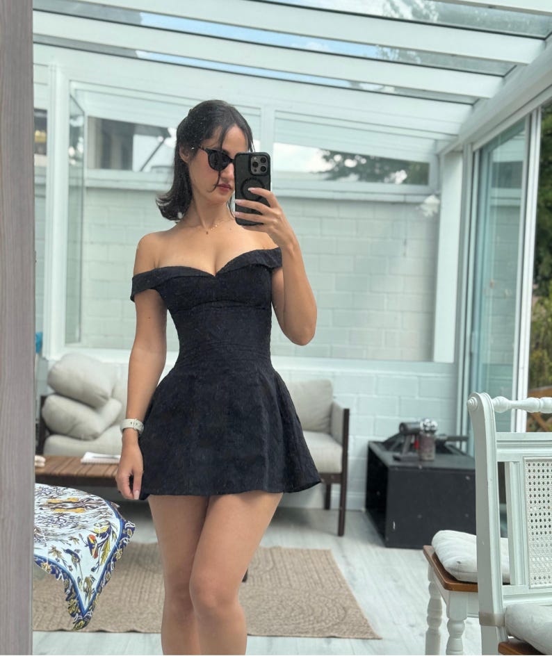 May include: A black off-the-shoulder dress with a fitted bodice and flared skirt. The dress has a textured pattern and is worn with sunglasses. The image is a selfie taken in front of a mirror.