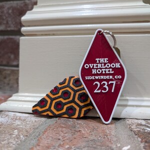 Pu&ograve; includere: Portachiavi a forma di diamante rosso con il testo "The Overlook Hotel" e "Sidewinder, CO" e il numero "237". Il portachiavi &egrave; accanto a un portachiavi con motivo nero e arancione.