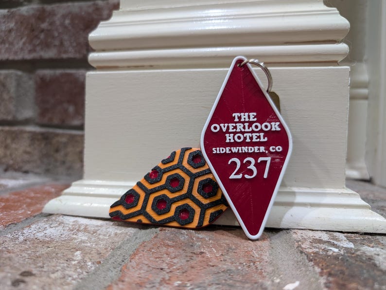Pu&ograve; includere: Portachiavi a forma di diamante rosso con il testo "The Overlook Hotel" e "Sidewinder, CO" e il numero "237". Il portachiavi &egrave; accanto a un portachiavi con motivo nero e arancione.