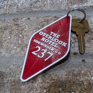 Pu&ograve; includere: Portachiavi rosso a forma di diamante con testo bianco che recita "The Overlook Hotel Sidewinder Co. 237".