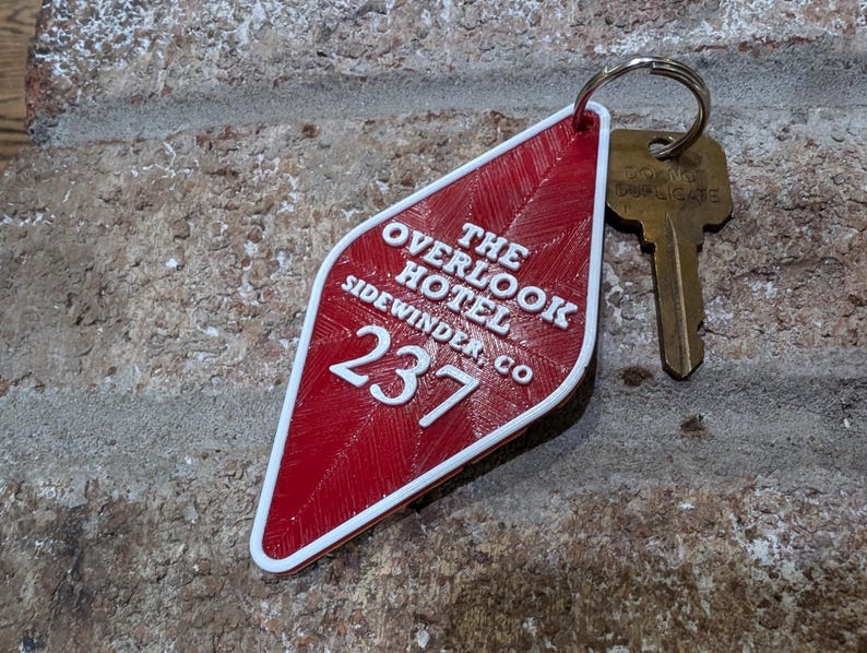 Pu&ograve; includere: Portachiavi rosso a forma di diamante con testo bianco che recita "The Overlook Hotel Sidewinder Co. 237".
