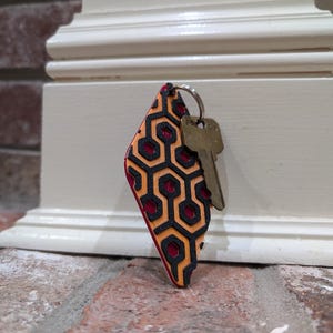 Pu&ograve; includere: Un portachiavi a forma di diamante rosso e nero con un motivo geometrico. Il portachiavi ha un anello argentato e due chiavi attaccate.