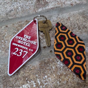 Pu&ograve; includere: Un portachiavi a forma di diamante rosso e bianco con il testo "The Overlook Hotel, Sidewinder, CO 237". Il portachiavi &egrave; attaccato a una chiave di ottone con un anello. &Egrave; visibile anche un secondo portachiavi a forma di diamante con un motivo geometrico nero e arancione.