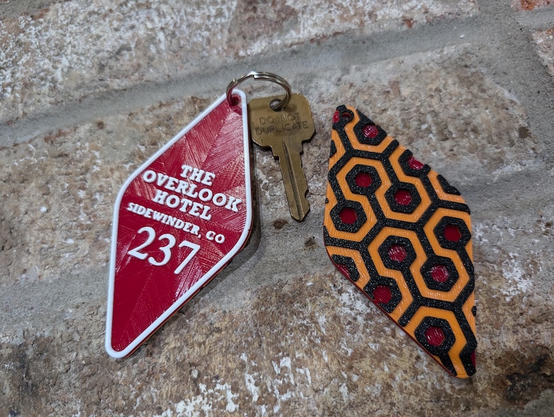 Pu&ograve; includere: Un portachiavi a forma di diamante rosso e bianco con il testo "The Overlook Hotel, Sidewinder, CO 237". Il portachiavi &egrave; attaccato a una chiave di ottone con un anello. &Egrave; visibile anche un secondo portachiavi a forma di diamante con un motivo geometrico nero e arancione.