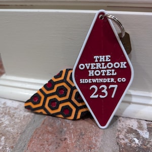Pu&ograve; includere: Un'etichetta per chiavi a forma di rombo rosso con il testo "The Overlook Hotel, Sidewinder, CO 237" in bianco. L'etichetta per chiavi &egrave; attaccata a un portachiavi argentato.