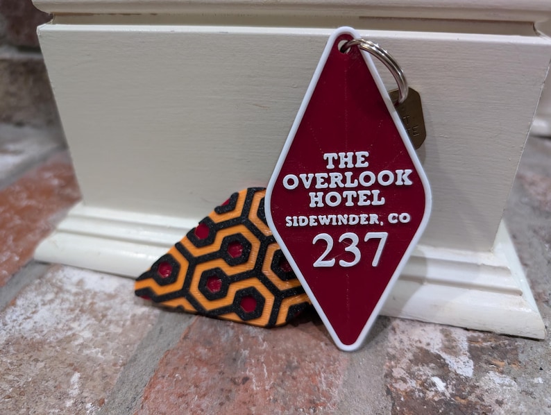 Pu&ograve; includere: Un'etichetta per chiavi a forma di rombo rosso con il testo "The Overlook Hotel, Sidewinder, CO 237" in bianco. L'etichetta per chiavi &egrave; attaccata a un portachiavi argentato.