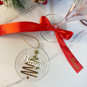 Può includere: Un ornamento natalizio rotondo e trasparente con il nome "Biagio" in scrittura bianca e un disegno di albero di Natale dorato. Un nastro rosso è legato attorno a un bicchiere di champagne, con il nome "Biagio" stampato sul nastro. Decorazioni festive.