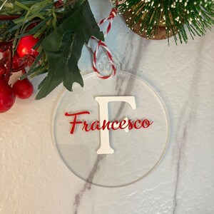 Può includere: Un ornamento natalizio rotondo e trasparente con il nome "Francesco" in corsivo rosso e una "F" bianca. L'ornamento è appeso a un cordino rosso e bianco. Sullo sfondo, vegetazione decorativa e un piccolo albero di Natale.