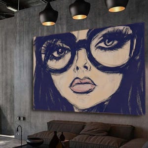 May include: A large canvas art print featuring a stylized illustration of a woman's face. The artwork showcases a close-up of the face, with prominent dark-rimmed glasses, long eyelashes, and full lips. The color palette includes shades of blue, beige, and pink.