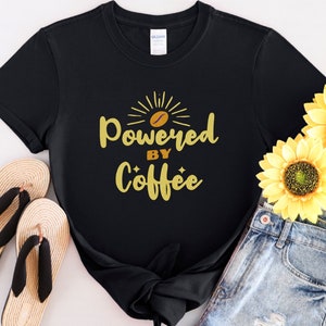 Puede incluir: Camiseta negra con un gráfico dorado que dice "Powered by Coffee" con un grano de café y un diseño de sol.