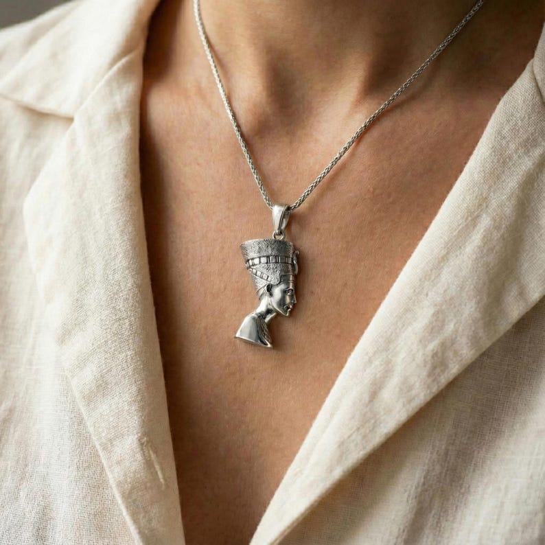 A silver pendant necklace with a pendant depicting a female profile, possibly a queen or princess, is worn around the neck of a person in a white shirt.