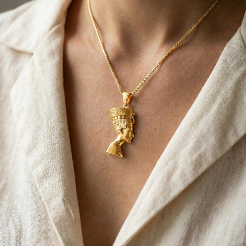 A gold-colored pendant necklace with a pendant depicting a female figure, possibly a queen or princess, is worn around the neck of a person in a white shirt.
