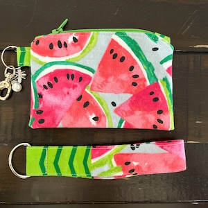 May include: A watermelon-themed accessory set featuring a small zippered pouch and a wristlet key fob. The pouch and wristlet are adorned with a vibrant pattern of watermelon slices in shades of red, green, and black on a light blue background.