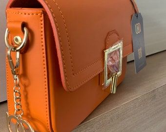 May include: A bright orange leather crossbody bag with a gold clasp and chain strap. The bag has a small pocket on the front and a tag with the text "BORSEIRE ARTIGIANE" on the side.