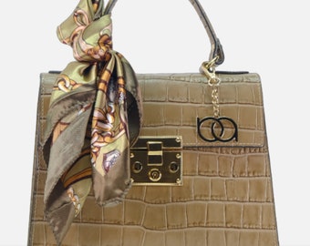 May include: A beige crocodile embossed leather handbag with a gold lock and chain detail. The bag has a top handle and comes with a matching scarf.