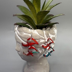 May include: A pair of abstract dangle earrings with a red, white, and teal design. The earrings are suspended from silver-colored hooks. The earrings are displayed in front of a white bust planter with a green succulent plant.