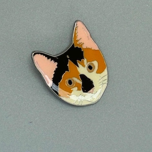 May include: A cat-shaped enamel pin with a silver border. The pin features a calico cat design with black, orange, and white colors. The cat's ears are pink. The pin is a cute accessory for cat lovers.