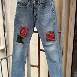 Może przedstawiać: Dżinsy z niebieskiego denimu z czerwonymi i czarnymi łatkami w kratę na przodzie każdej nogawki.