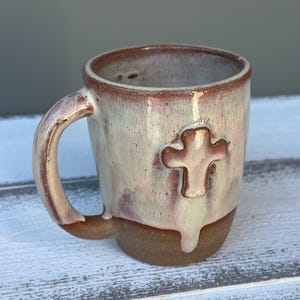 May include: A handmade ceramic mug with a cross design. The mug features a brown base, a cream-colored upper section with a cross, and a brown handle. The mug has a rustic, textured appearance.
