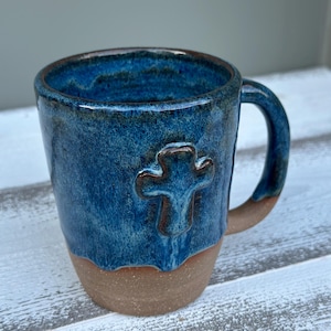 May include: A handmade ceramic mug with a blue glaze and a brown base. The mug features a raised cross design on the front. The handle is also blue. This unique mug is perfect for coffee or tea.