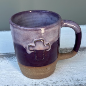 May include: Handmade ceramic mug with a cross design. The mug features a two-tone glaze, with a deep purple base and a lighter purple top. The handle and cross are also purple. The bottom of the mug is a natural tan color.