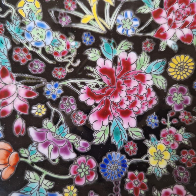 May include: Close-up of a black ceramic plate with a colorful floral design. The flowers are in shades of pink, red, blue, yellow, and purple, with green leaves. The design is intricate and detailed, with a glossy finish.