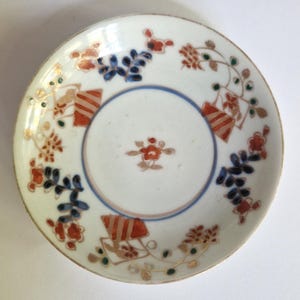 May include: A round white ceramic saucer featuring a central red floral motif surrounded by a blue circular line. The rim is decorated with repeating patterns of red and blue floral sprays and stylized orange and white striped rectangular shapes.