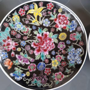 May include: A decorative plate with a black background and a vibrant floral design. The flowers are in shades of red, pink, blue, yellow, and purple, with green leaves. The plate has a white rim.
