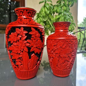 Peut inclure: Deux vases décoratifs rouges avec des sculptures florales complexes. Le plus grand vase a un fond noir, tandis que le plus petit est entièrement rouge. Les deux vases présentent un design sculpté et une forme classique.