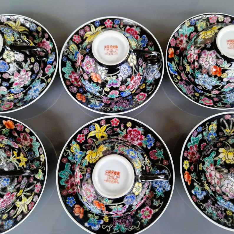 May include: Set of six black teacups and saucers with colorful floral designs. Each cup and saucer features a black background with a vibrant array of pink, yellow, blue, and orange flowers. The cups have black handles and the saucers have a white rim.