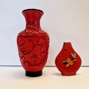 May include: Two red Chinese cinnabar vases with intricate carved floral designs. The larger vase has a black base and the smaller vase has a gold top.
