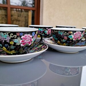 May include: Set of six black teacups and saucers with floral designs. Each cup features a pink peony and colorful accents. The saucers and cups have a white rim and are set on a reflective surface.