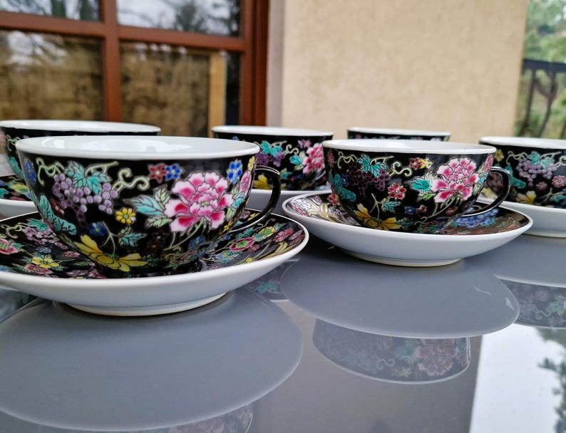 May include: Set of six black teacups and saucers with floral designs. Each cup features a pink peony and colorful accents. The saucers and cups have a white rim and are set on a reflective surface.