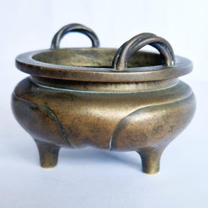 May include: A bronze incense burner with a round body, three short legs, and two arched handles. The rim of the burner is wide, and the design is simple and elegant. The metal has a slightly aged appearance.