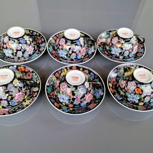 May include: Six porcelain teacups and saucers. The cups and saucers are black with a floral design in shades of pink, blue, yellow, and orange. Each cup has a black handle and a white interior. The saucers have a white rim.