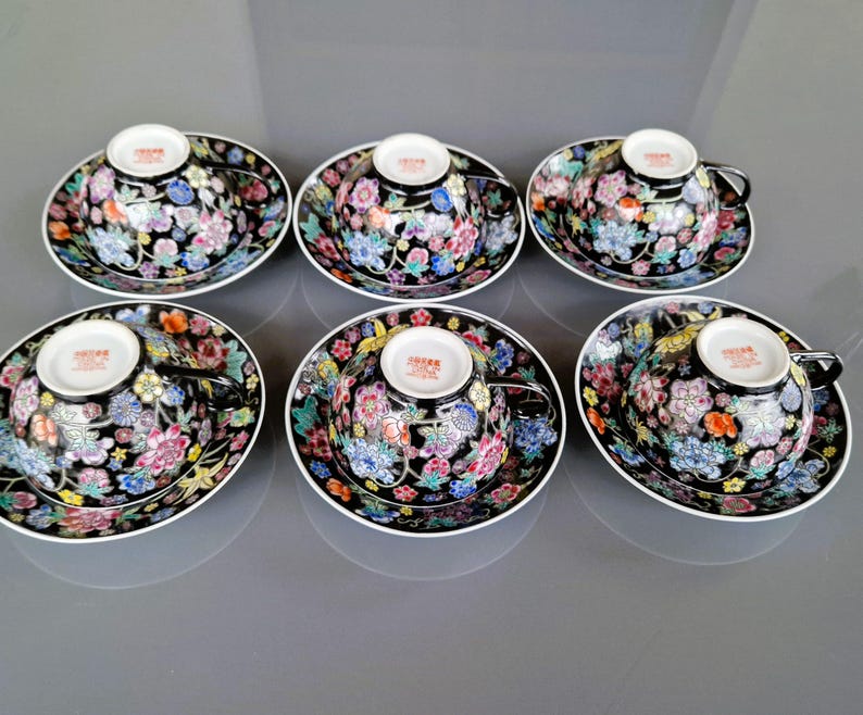 May include: Six porcelain teacups and saucers. The cups and saucers are black with a floral design in shades of pink, blue, yellow, and orange. Each cup has a black handle and a white interior. The saucers have a white rim.