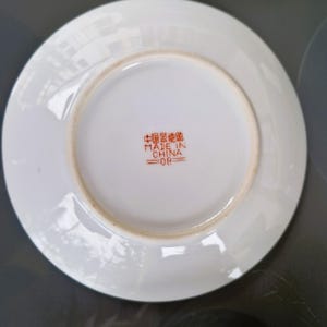 May include: Underside of a white ceramic saucer with a slightly raised rim. The center features red Chinese characters and the text "MADE IN CHINA =08". The saucer has a light brown ring near the edge.