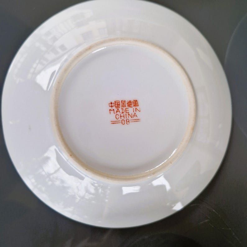May include: Underside of a white ceramic saucer with a slightly raised rim. The center features red Chinese characters and the text "MADE IN CHINA =08". The saucer has a light brown ring near the edge.