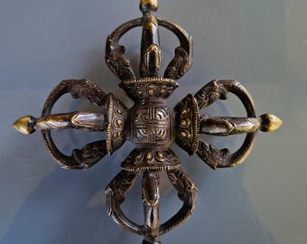 Può includere: Una ruota di preghiera tibetana in bronzo con dettagli intricati e una finitura dorata. La ruota ha la forma di una croce con quattro braccia, ciascuna terminante con una punta. Il centro della ruota è decorato con un disegno circolare.