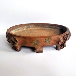 Może przedstawiać: Owalna doniczka ceramiczna do bonsai w kolorze brązowym o teksturowanej powierzchni przypominającej korę drzewa. Doniczka posiada cztery krótkie, solidne nóżki oraz dwa zielone znaki kaligraficzne wygrawerowane na przedniej stronie.