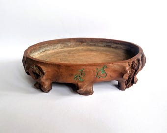 May include: An oval-shaped brown ceramic bonsai pot with a textured, bark-like exterior finish. The pot features four short, sturdy feet and two green calligraphic characters engraved on the front side, showcasing a natural, rustic appearance.