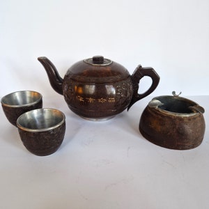 May include: A brown wooden tea set comprising a teapot, two cups, and an ashtray. The teapot and cups feature intricate carvings and gold lettering. The ashtray is also carved and has a metal insert.