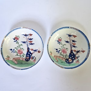 May include: Two white porcelain bowls with blue and red floral designs. The bowls have a green and white landscape scene at the bottom.
